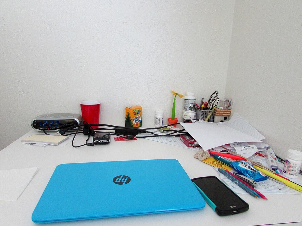 My desk with various paraphernalia, namely a blue laptop, box of Crayola crayons, two point-and-shoot cameras, papers, pens