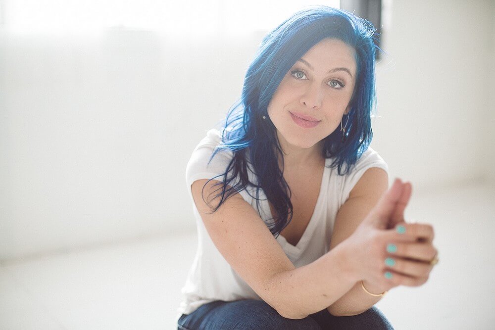 Photo of Alexandra sitting in a white room. Her hair and pants are blue, her shirt white, and her nails a light blue.