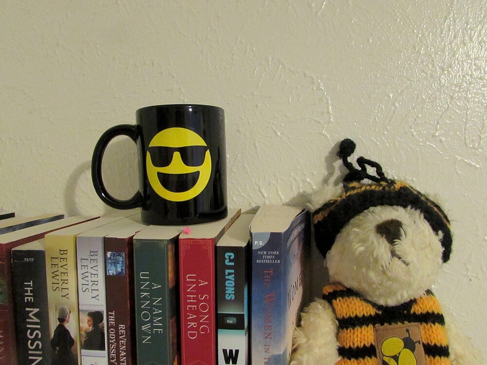 Sunglasses emoji mug on top of books on a shelf, next to a white teddy bear dressed in a bee costume