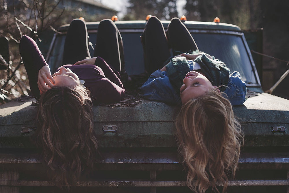 Two girls laying on their backs on the hood of an old, rusty truck, laughing; their wavy hair hangs free off the edge of the hood. They are in long-sleeved tops and long pants, and one is wearing a scarf
