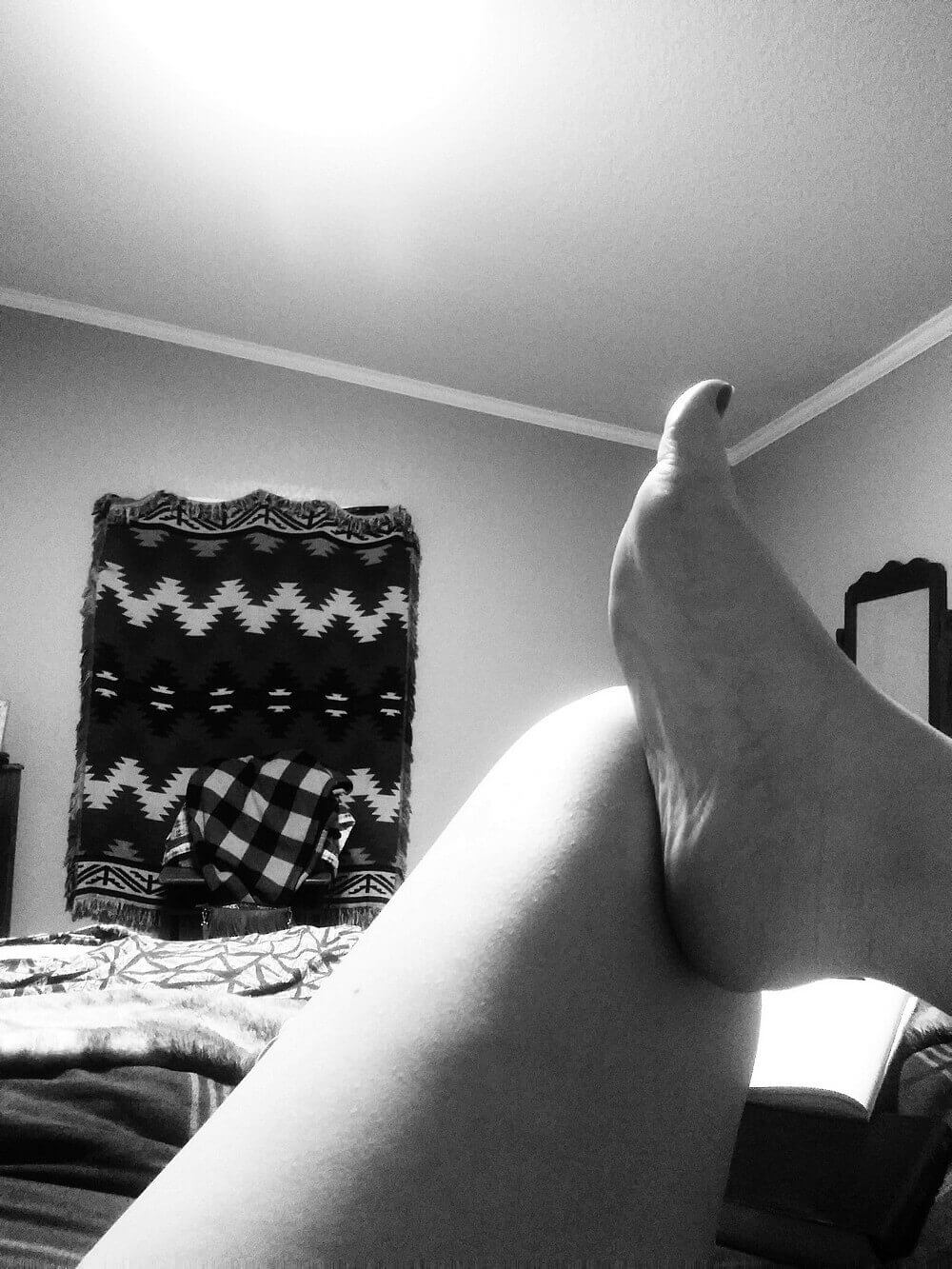 Bare leg and foot on bed, warm glow created by ceiling light; textured pattern curtain in background; black and white