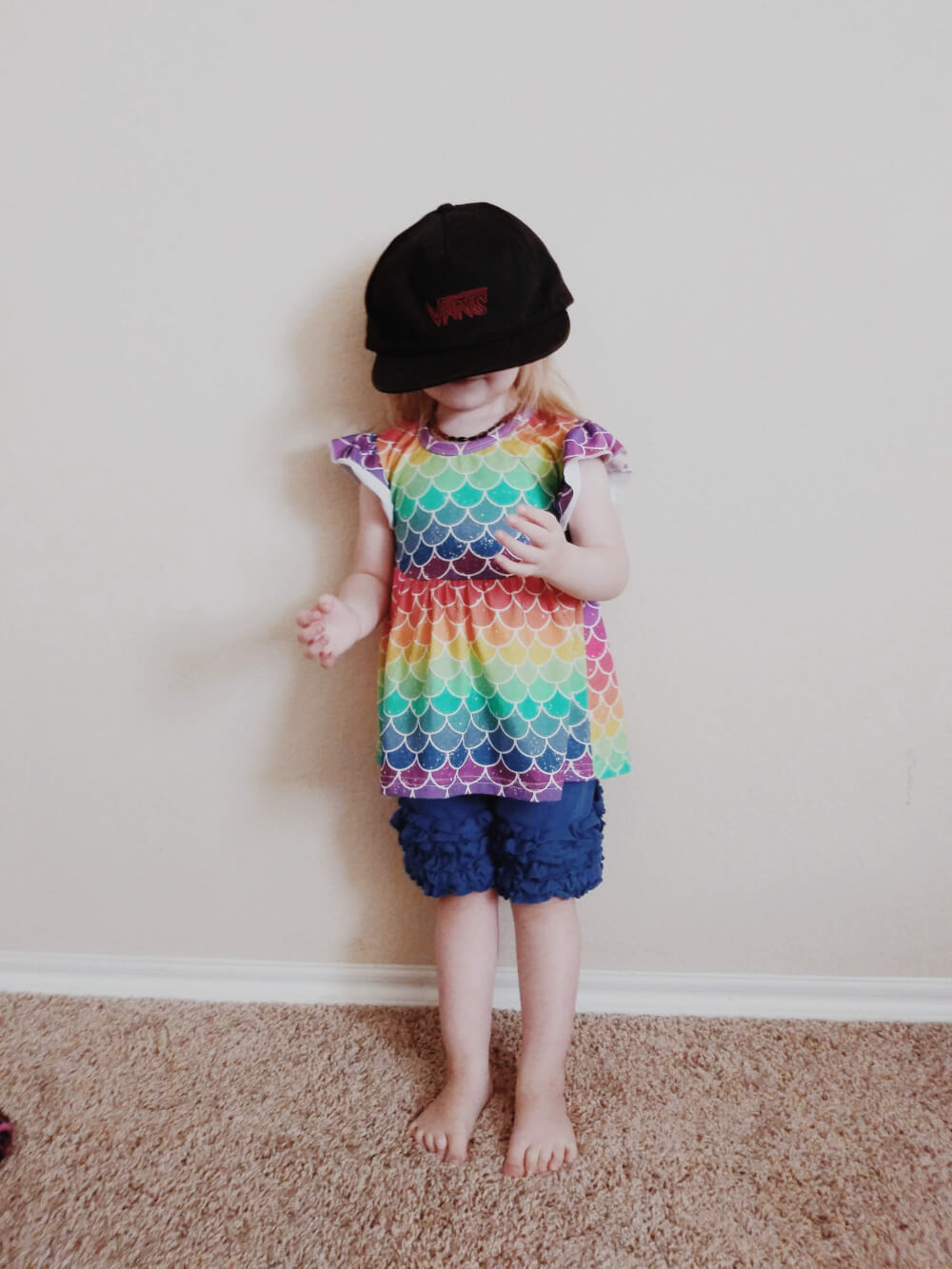 Toddler in rainbow scales outfit wearing a cap too big