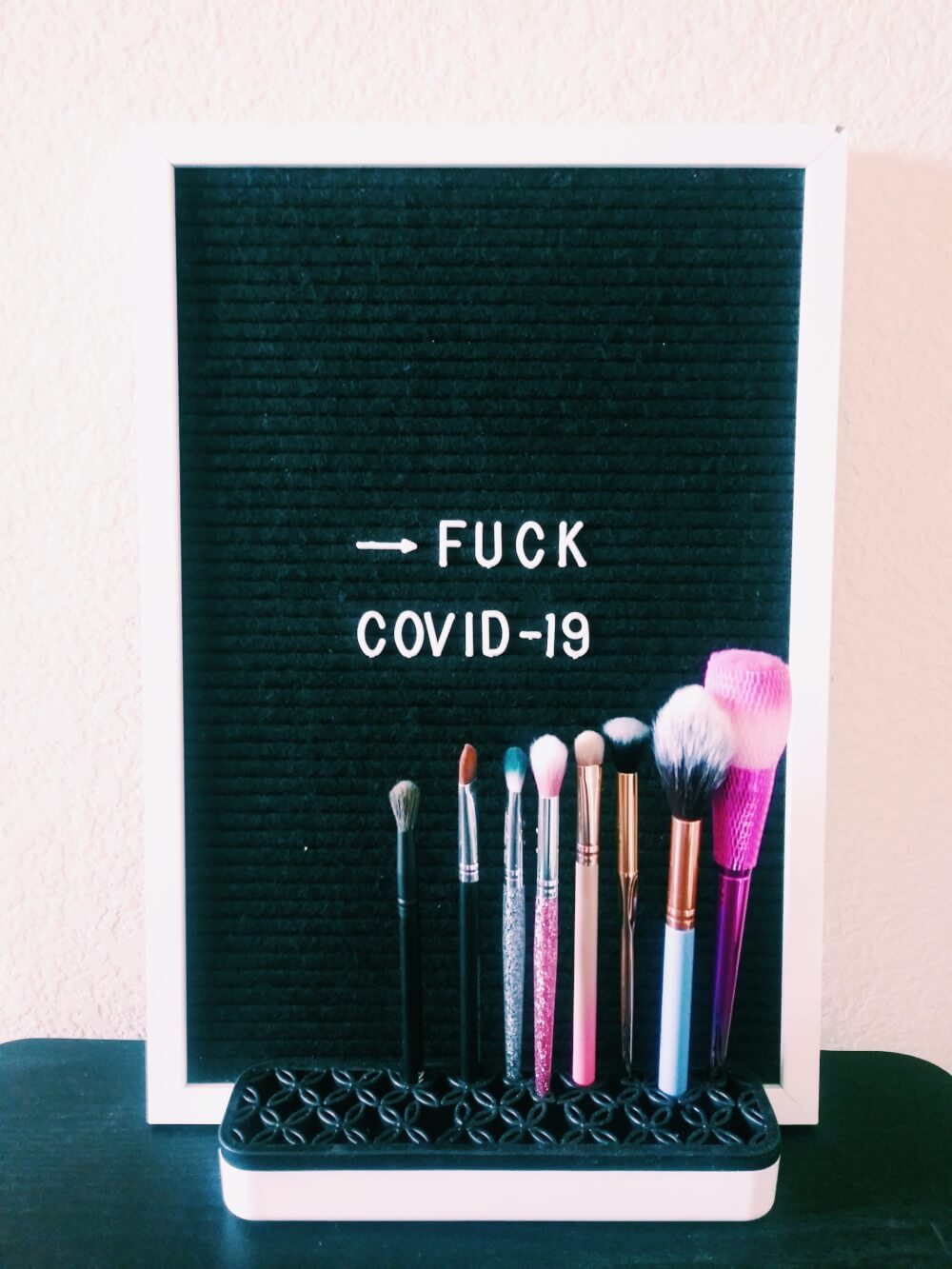 Letterboard saying "FUCK COVID-19" behind a row of makeup brushes