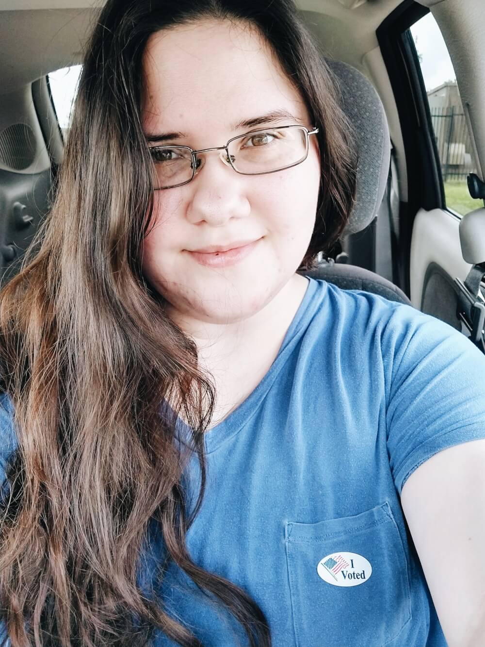 Car selfie in blue top with "I voted" sticker and wavy hair