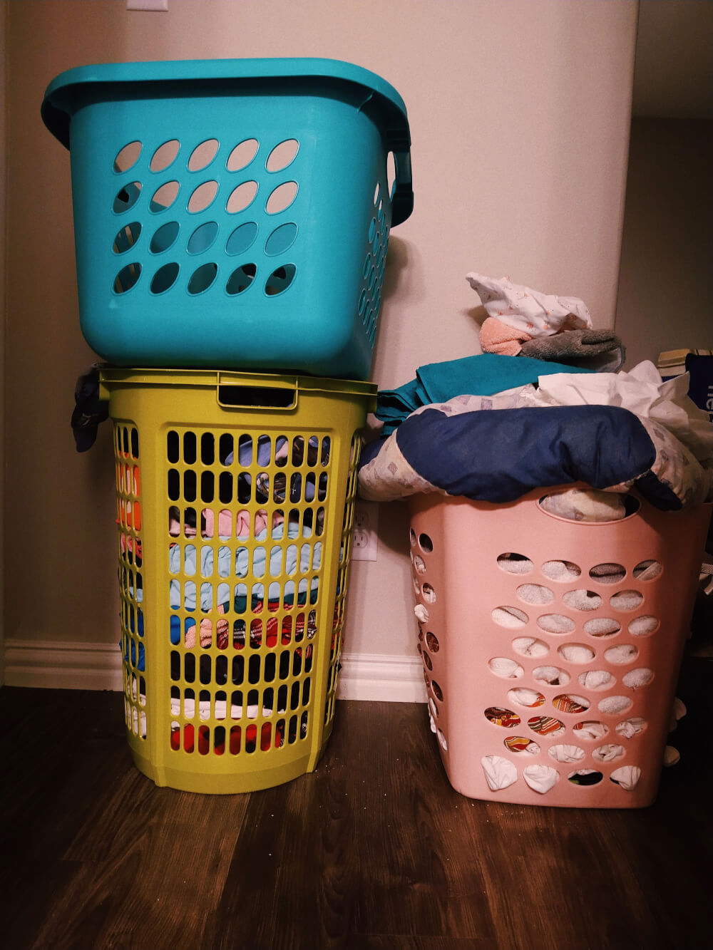 Empty teal basket atop lime green basket full of dirty clothes, next to blush pink basket overflowing with dirty laundry
