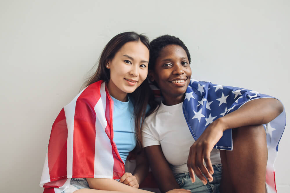 An asian woman and a Black woman both smiling while sitting close together, with the US flag wrapped around their shoulders