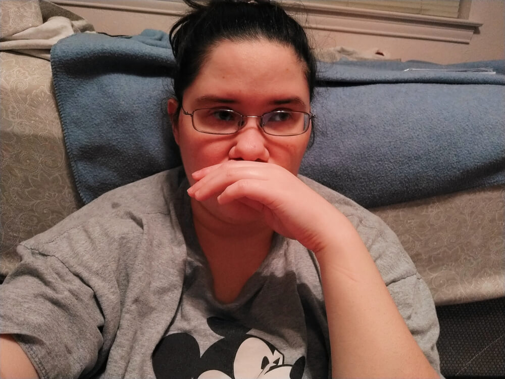 me, distraight, sitting on the floor in front of a bed, eyes looking off to the side, hand between upper lip and nose, wearing a grey Mickey Mouse shirt